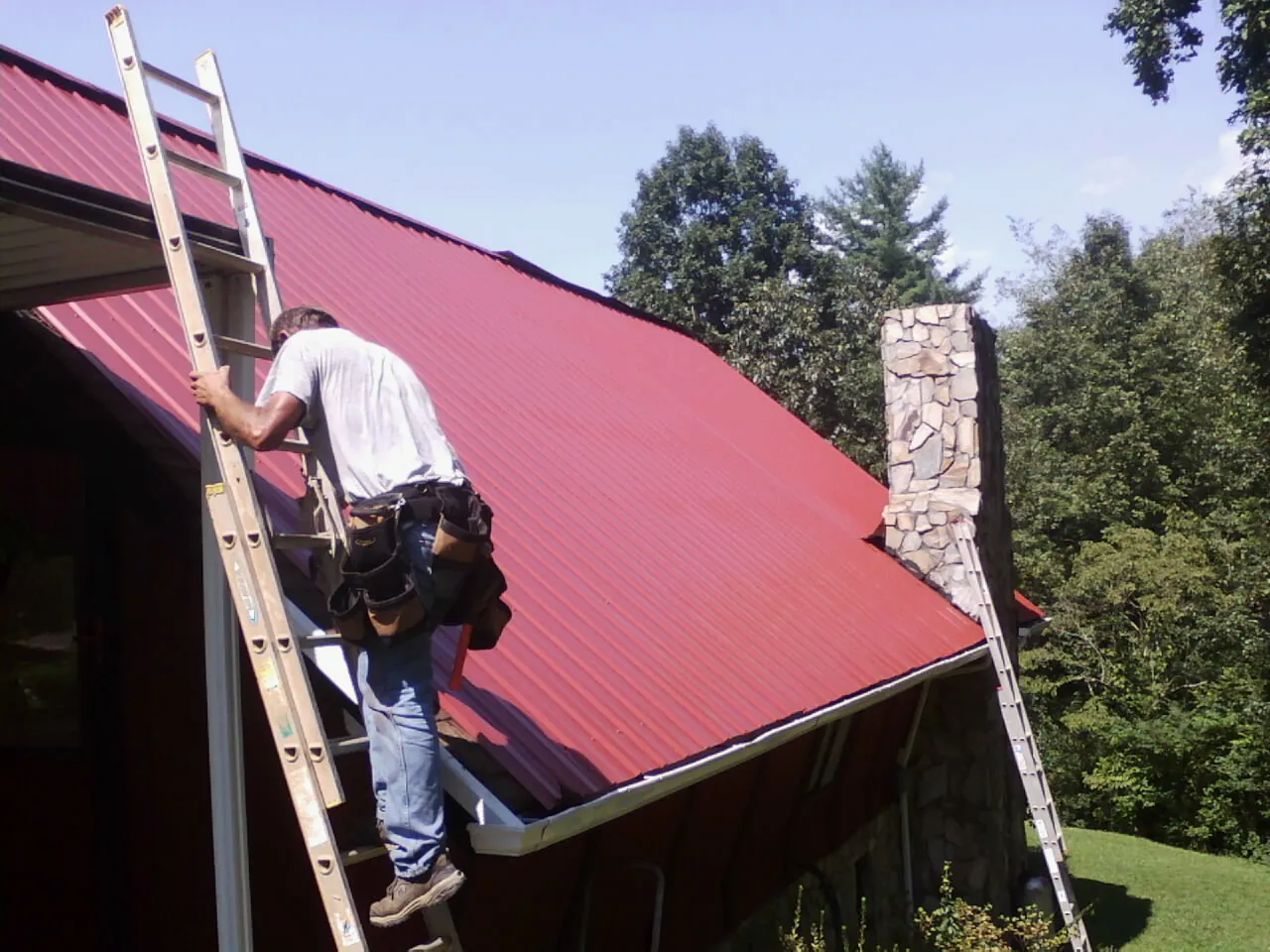 Roofer on a ladder beside a red metal roof and stone chimney in Swanlake